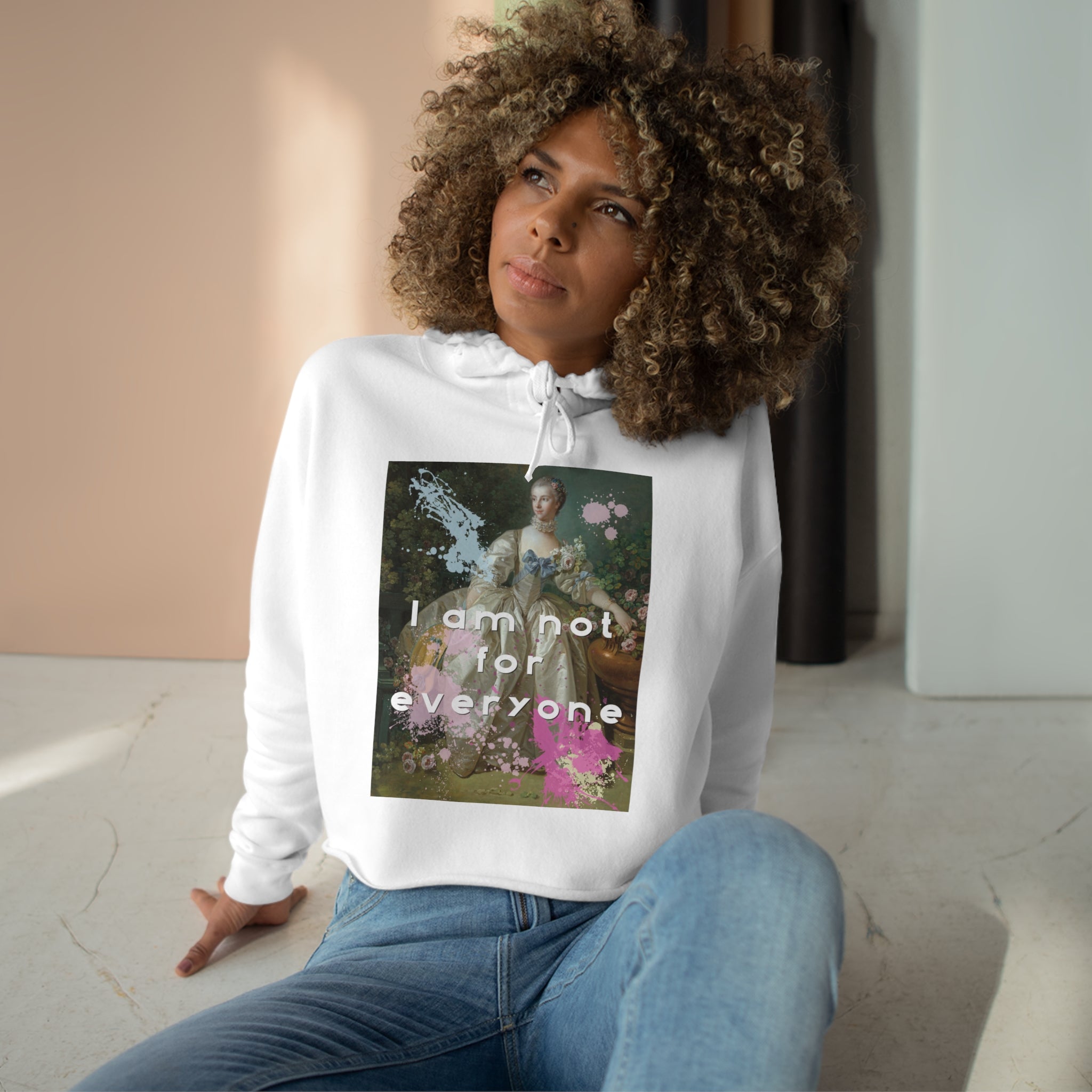 Lifestyle image of the cropped hoodie in white features and altered piece of classical art on the front. The woman has paired this with classic blue jeans. The altered piece is the original Madame Bergeret from 1766, a high society girl in a off-white voluminous dress surrounded by flowers. We have modernized the piece by adding bold and bright paint splashes in yellow, light blue, and hot pink and the phrase,” I am not for everyone”